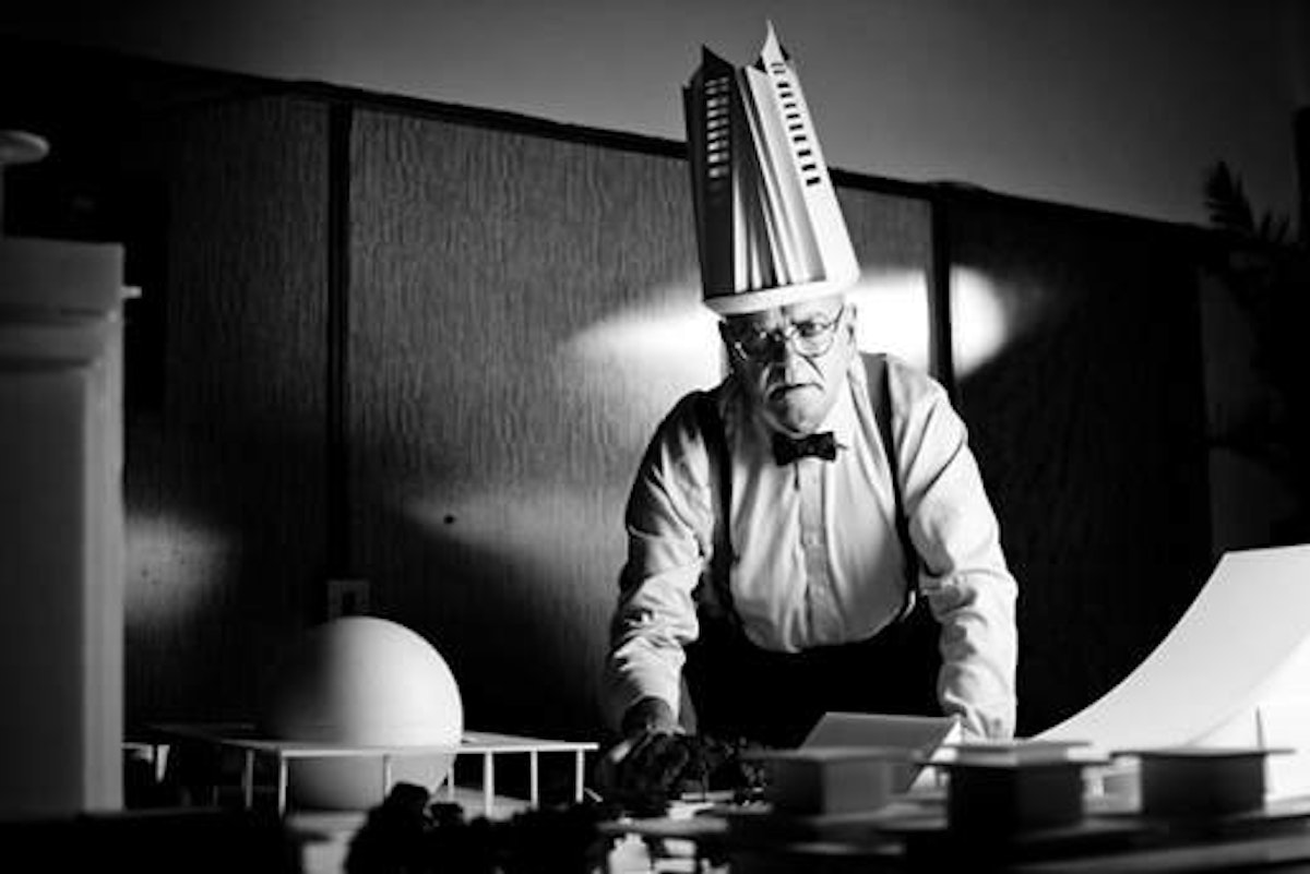 A older man with a building shaped hat leans over a desk